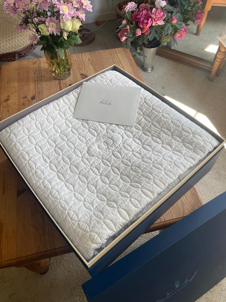Matelas matelassé blanc dans une boîte cadeau bleu, posé sur une table en bois, avec des bouquets de fleurs au fond.